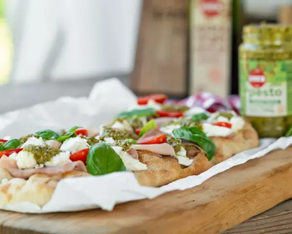 Długi placek z mozzarellą, pomidorkami i bazylią, polany zielonym pesto. Obok słoiczek z pesto.