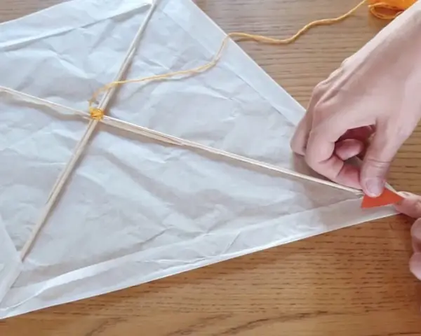 Mains fabriquant un cerf-volant simple en papier blanc et bâtons de bois, avec fil jaune.