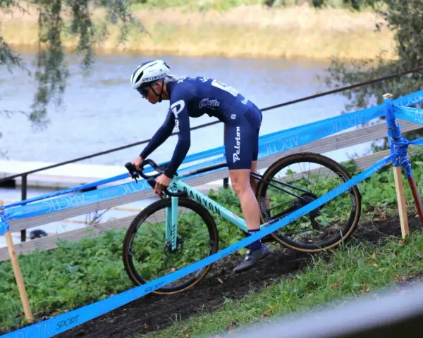 Cyclo cross racer in blue kit navigates a muddy course beside a lake, marked by blue tape.