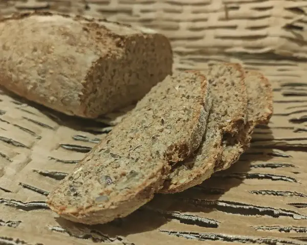Chleb orkiszowy, pokrojony w kromki, z widocznymi ziarnami, leży na falistej tekturze.