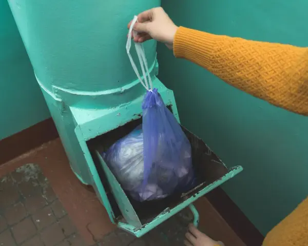 Ręka w żółtym swetrze wkłada worek ze śmieciami do metalowego zsypu w bloku.