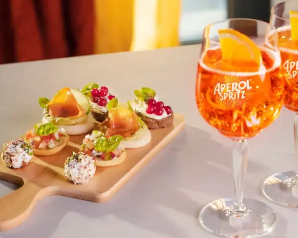Zwei Aperol Spritz Gläser mit Orangenscheiben und eine Holzplatte mit verschiedenen Häppchen. Perfekt für einen Aperitif!