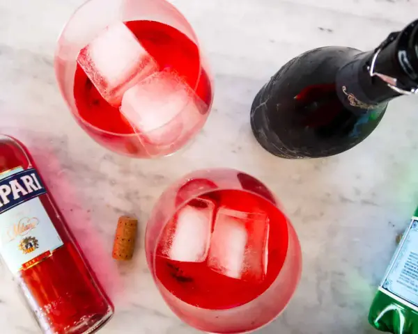 Dwa orzeźwiające Campari Spritz z lodem, butelka Campari i butelka prosecco na marmurowym blacie.