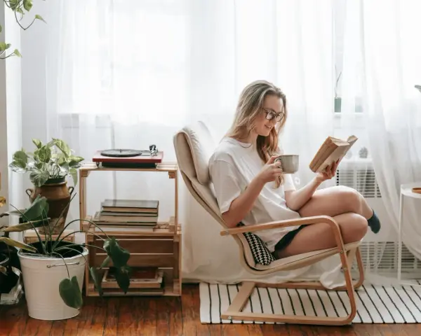 Mujer con gafas disfruta de un libro y una taza en su rincón de lectura, rodeada de plantas y luz natural.