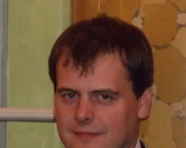 Tomasz Bartoś, zamyślony, w eleganckim stroju, na tle ozdobnej ściany.