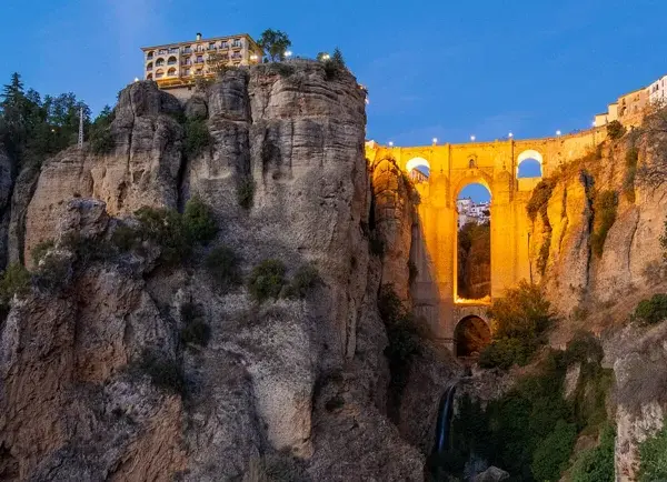 A dramatic **Ronda bridge view point** at dusk, with the illuminated bridge spanning a deep gorge and buildings perched on the cliff edge.