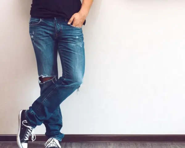 Mężczyzna w modnych jeansach z dziurami na kolanie, czarnych trampkach i czarnej koszulce, opiera się o ścianę.
