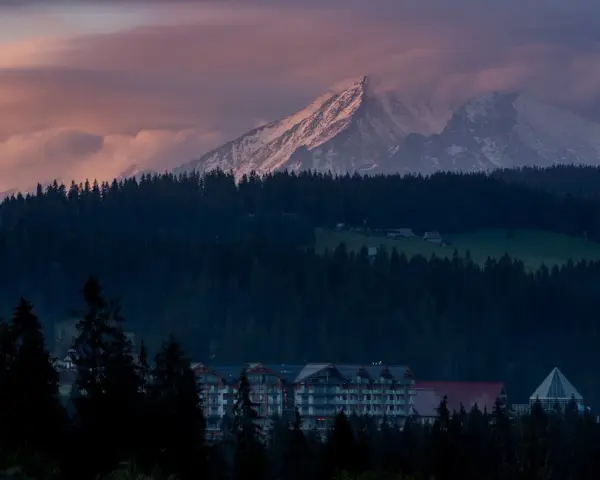 Górski krajobraz o zachodzie słońca z ośnieżonymi szczytami, lasem i budynkami hotelowymi.