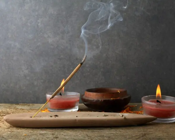 Un incienso humeante se posa sobre un soporte de arcilla, rodeado de velas rojas y un cuenco de madera. Perfectos inciensos para limpiar energías y crear un ambiente de paz.