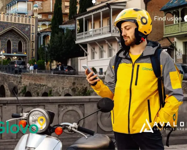 Kurier Glovo sprawdza telefon, gotowy do pracy w blokach czasowych. Na skuterze widnieje logo.
