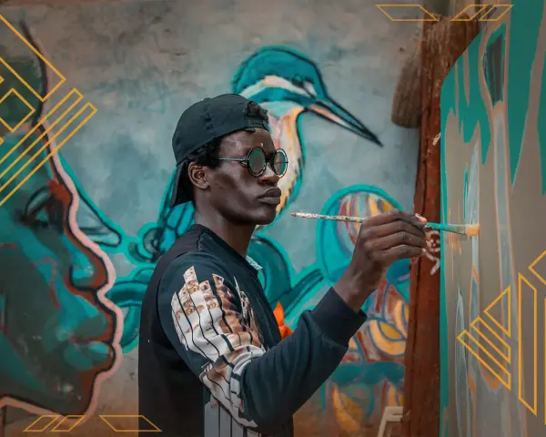 Un artiste au style d'art peinture, casquette et lunettes rondes, peint une fresque murale colorée représentant un visage et un oiseau.