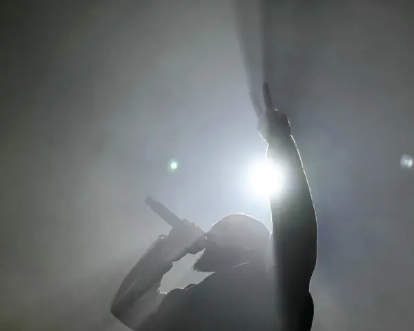Un rappeur, micro en main, lève un doigt en signe de paix. Lumière intense, fumée, ambiance d'un exposé sur le rap.