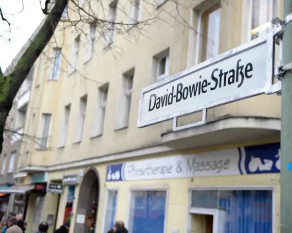 Straßenschild "David-Bowie-Straße" in Berlin. Ein Baum und ein Gebäude mit Geschäften im Hintergrund.