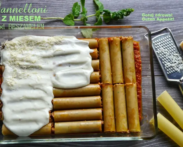 Cannelloni z mięsem mielonym i sosem beszamelowym w naczyniu żaroodpornym, gotowe do pieczenia. Obok tarka i kawałek parmezanu.