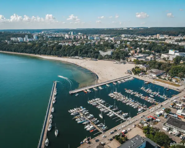 Widok z lotu ptaka na plażę miejską w Gdyni, z zatoką pełną jachtów i łodzi, oraz miastem w tle.