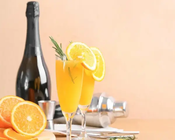 Dwa kieliszki z pomarańczowym napojem, ozdobione plasterkami pomarańczy i rozmarynem. Obok butelka szampana, shaker i świeże pomarańcze. Mimosa czy Bellini?
