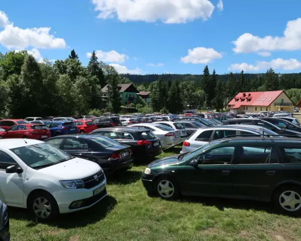 Tłum samochodów na parkingu przy Karłowie Szczeliniec. Słoneczny dzień, chmury na niebie, otoczenie lasu i budynków.