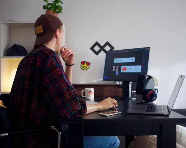 Młody mężczyzna w czapce i flanelowej koszuli pracuje przy komputerze. To typowy freelancer, który tworzy swoje projekty z domu.