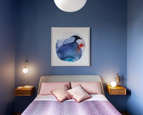 Camera da letto con pareti blu intenso e un quadro astratto. Cuscini rosa e coperta viola creano colori rilassanti.