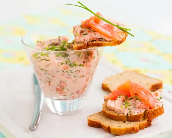Rillettes de saumon crémeuses dans un verre, garnies de fines herbes et servies sur des toasts avec des tranches de saumon fumé.