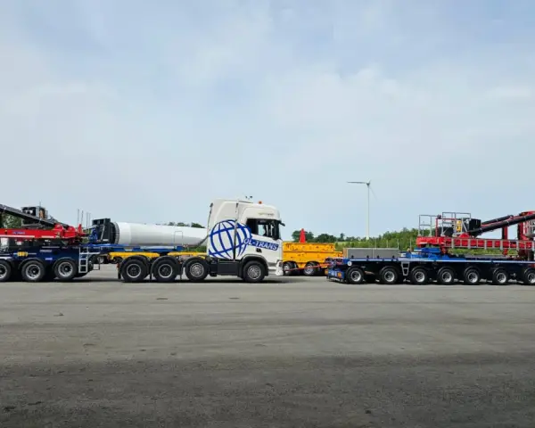 Ciężarówka z naczepą tandem przewozi element turbiny wiatrowej. Obok stoi inna naczepa z dźwigiem.