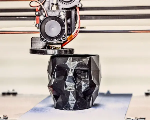 A 3D printer nozzle extrudes filament to create a faceted black skull planter, showcasing innovative 3D printing examples.