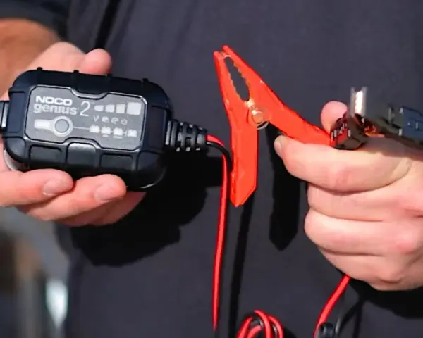 A person holds a NOCO Genius 2 trickle charger and its red clamp. This charger is designed to prevent damage, so no, a trickle charger can't ruin a battery when used correctly.