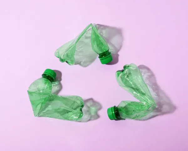 Trzy zielone, zgniecione butelki tworzą symbol recyklingu na różowym tle.