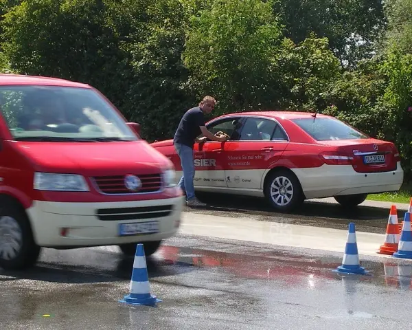 Mann reinigt Taxi. Ein roter Transporter fährt vorbei. Verkehrskegel markieren den Bereich. Möglicherweise eine Schulung der Berufsgenossenschaft Taxi.