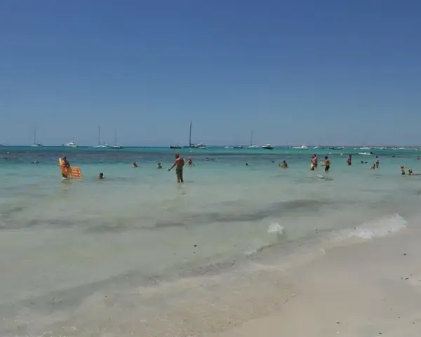 Słoneczny dzień na plaży Es Trenc. Ludzie kąpią się w turkusowej wodzie, a na horyzoncie widać żaglówki.