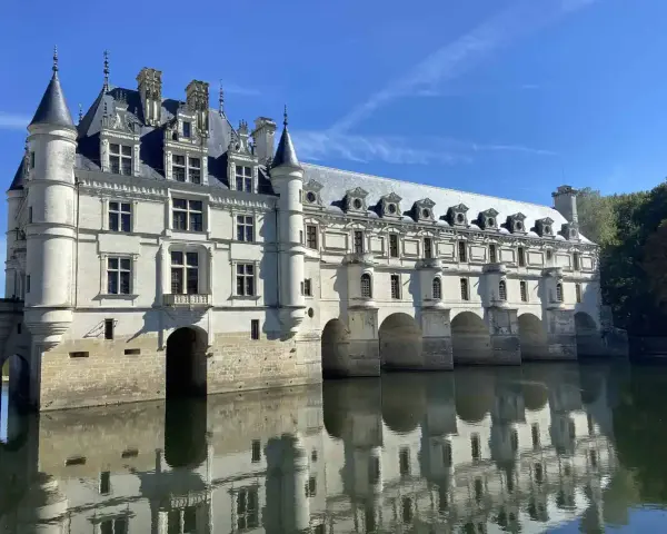 The stunning Chateau de Chenonceau, a white stone castle, gracefully spans the River Cher, its reflection shimmering on the water's surface under a clear blue sky.
