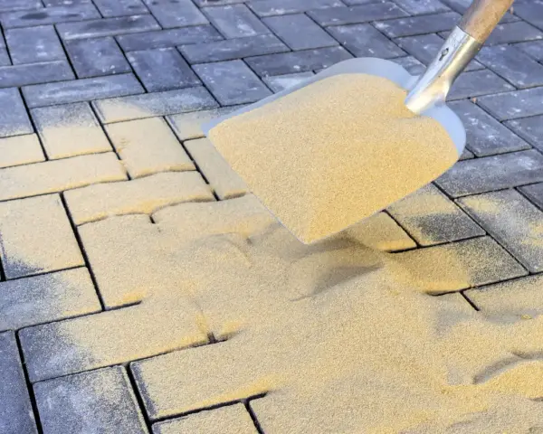 Schaufel mit Sand zum Terrassenplatten verfugen. Sand rieselt auf graue Pflastersteine.