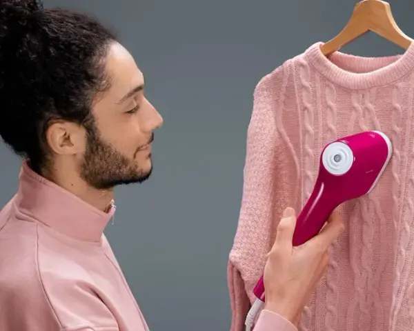 Man using a pink handheld steamer on a pink cable-knit sweater hanging on a wooden hanger.