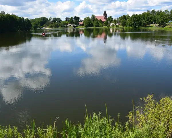 Spokojne jezioro na Kaszubach, odbijające chmury. W oddali widać łódkę i kościół. Idealne miejsce na wakacje.