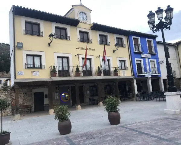El Ayuntamiento de Tajuña, con su balcón y reloj, preside la plaza. Edificios de colores y farolas completan la escena.