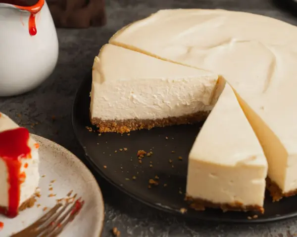 Ein cremiger New York Cheesecake, das Original-Rezept, mit einem Stück auf einem Teller, beträufelt mit roter Soße.