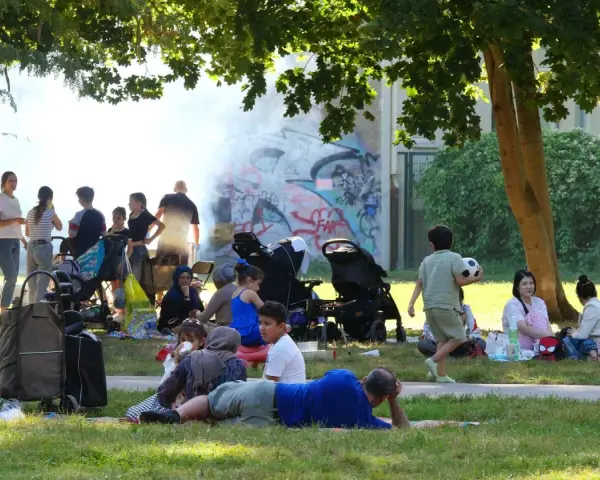 Familien genießen den Sommer auf einem Grillplatz in Berlin. Rauch steigt von einem Grill auf, während Kinder spielen und sich entspannen.