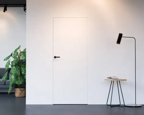 Białe drzwi ukryte w ścianie z czarną klamką, obok stojąca lampa i stolik.