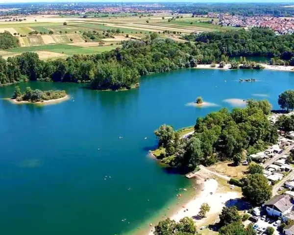 Der Mechtersheim See mit seiner türkisen Wasserfarbe, umgeben von grünen Bäumen und einem Campingplatz. Im Hintergrund erstrecken sich Felder und eine Stadt.