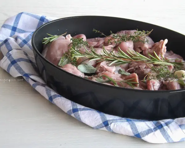 Pezzi di coniglio crudo pronti per essere cucinati al forno, profumati con rosmarino, salvia e aglio.