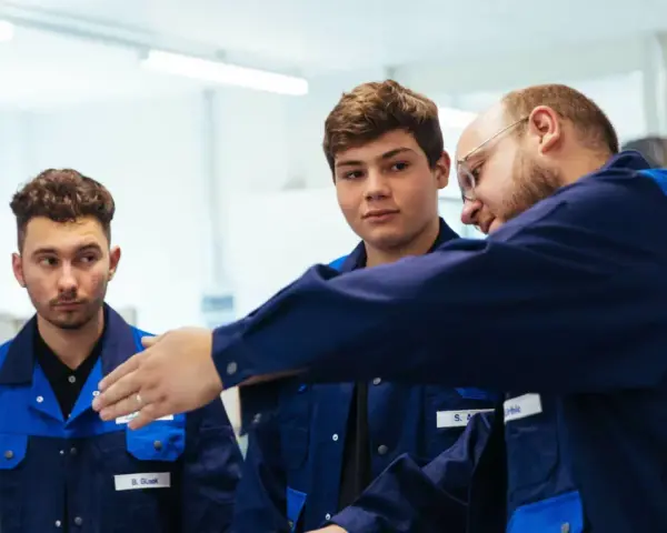 Drei junge Männer in blauen Arbeitskitteln lernen gemeinsam. Ein Ausbilder erklärt etwas, während die genz-Auszubildenden aufmerksam zuhören.