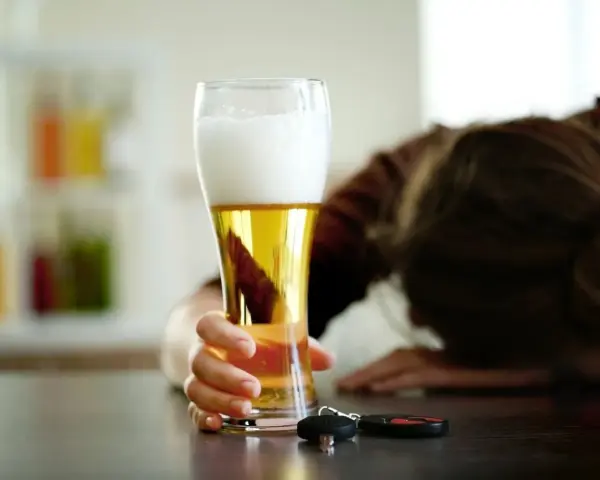 Ręka trzymająca piwo, obok kluczyki samochodowe. Osoba w tle pochylona.