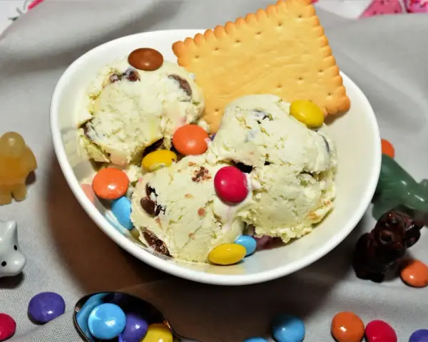Schüssel mit cremigem Eis, garniert mit bunten Smarties und einem Keks. Kleine Figuren schmücken die Szene.