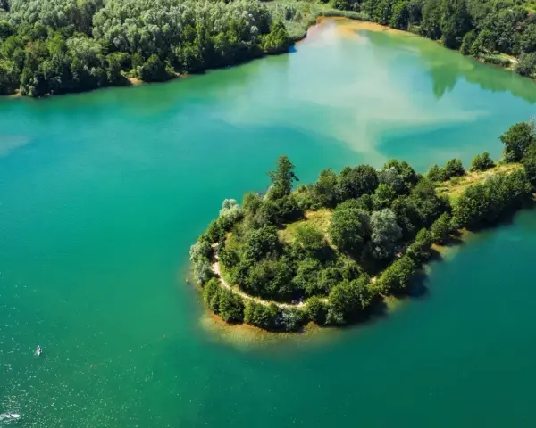 Türkisblaues Wasser umgibt eine grüne Insel mit einem Pfad am Niedernberger See. Ein Stand-Up-Paddler genießt die Ruhe.