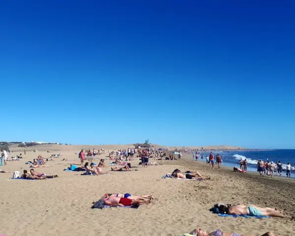 Tłum ludzi odpoczywa na słonecznej plaży. Woda jest spokojna, a niebo bezchmurne. Czy Gran Canaria jest bezpieczna? Tak, wygląda na to, że turyści czują się tu swobodnie.