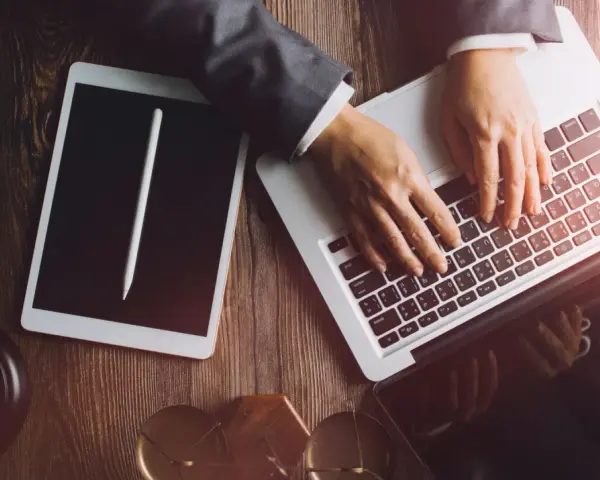 Dłonie na klawiaturze laptopa, obok tablet i młotek sędziowski. Profesjonalne narzędzia do pracy, być może związane z **taxat windykacja**.