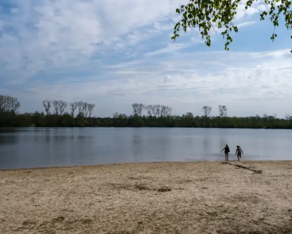 Zwei Kinder planschen im Wasser am Sandstrand des Dieksees in Lingen. Bäume säumen das Ufer unter einem bewölkten Himmel.