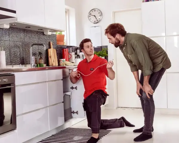 Tekniker installerar skalskydd larm under diskbänken i ett modernt kök. En man tittar på.