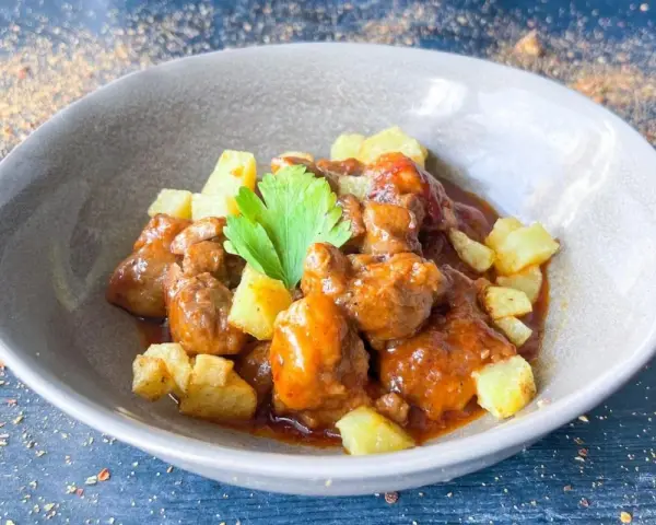 Delicioso cordero al chilindrón en un cuenco gris, acompañado de patatas fritas y una ramita de perejil.