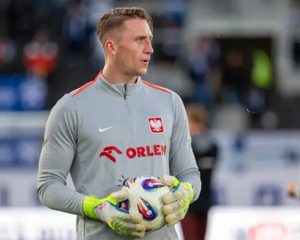Bramkarz w szarej bluzie z orłem i napisem ORLEN, trzymający piłkę. Kolejny z polscy piłkarze za granicą, gotowy do gry.